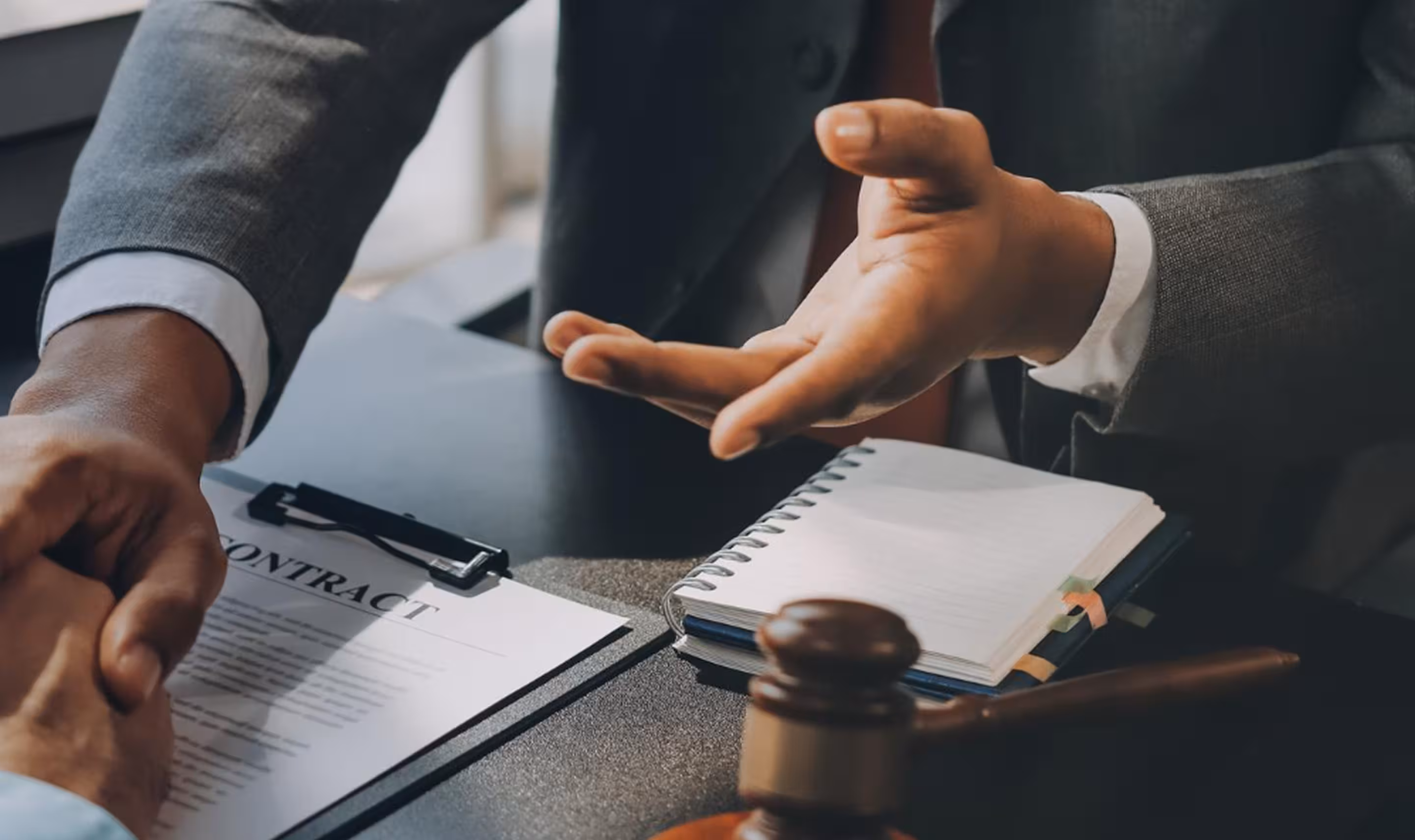 Two people shaking hands over a contract document with a gavel and notebook on the table.