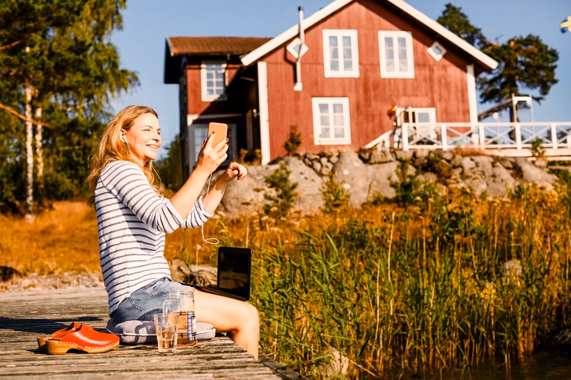 Kvinne sitter på en brygge med mobil og laptop foran en rød hytte