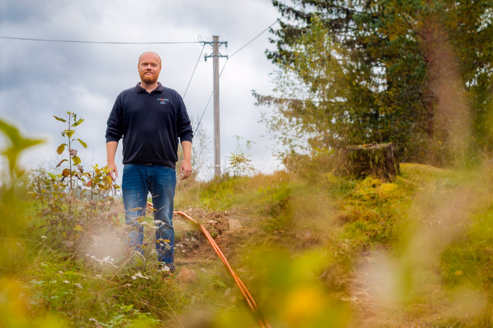 Altifiber-montør står i terrenget der ny fiberkabel legges ned, med skog og strømlinje bak