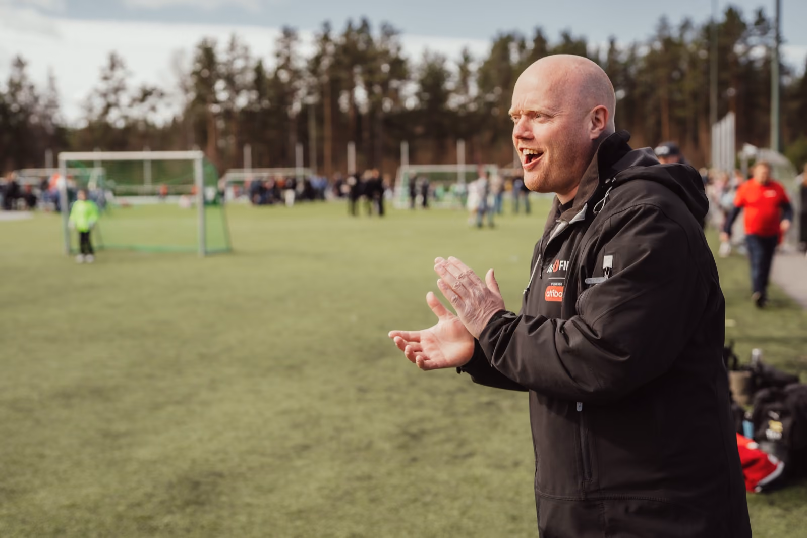 Altifiber-ansatt i jakke med logo som heier fram et lag på fotballbanen