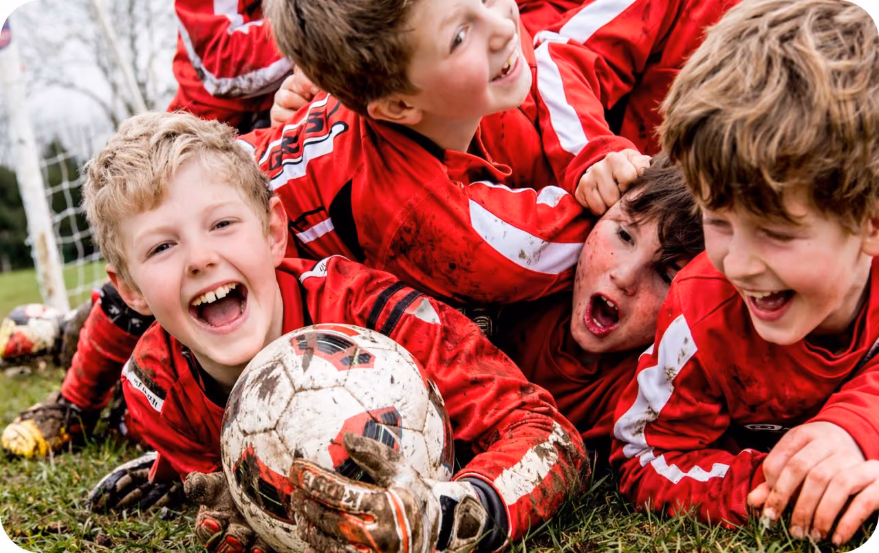 Barn i røde fotballdrakter ligger i gresset og ler sammen rundt en fotball