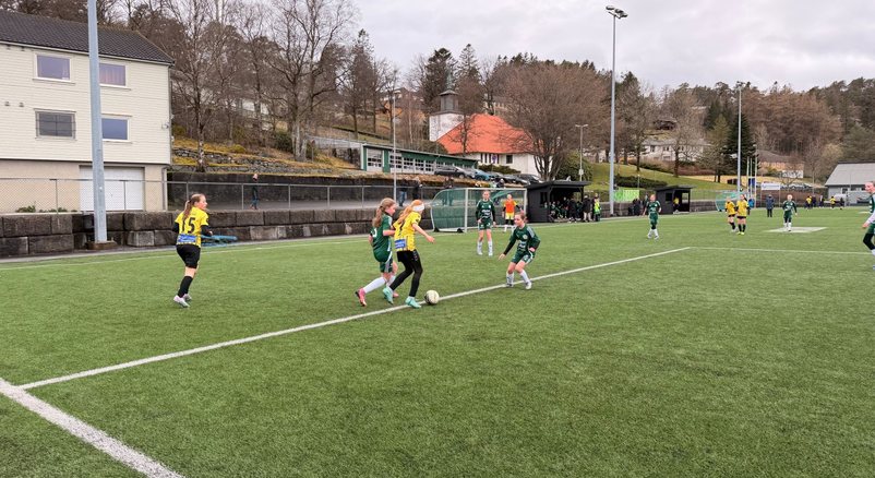 Jublende barn i røde fotballdrakter ligger tett sammen i gresset rundt en fotball.