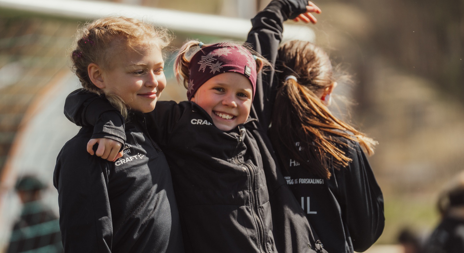 To jenter i svarte klubbjakker står tett sammen foran et fotballmål og ler