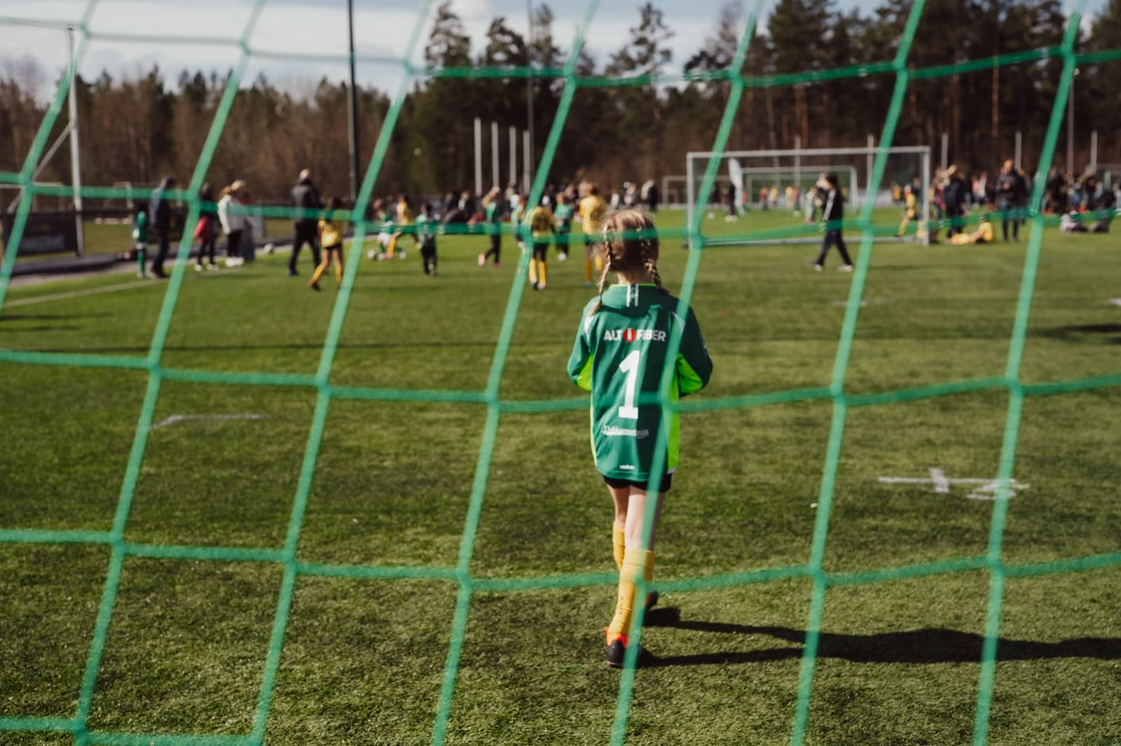 Barn i Altifiber-drakt på fotballbanen, sett gjennom målnettet