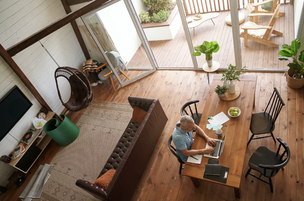 Top-down view of a man seated at a wooden dining table working on a laptop in a bright, modern living room with wooden floors, plants, a brown leather sofa, hanging wicker chair, and large glass doors opening to a balcony with outdoor chairs and cushions.