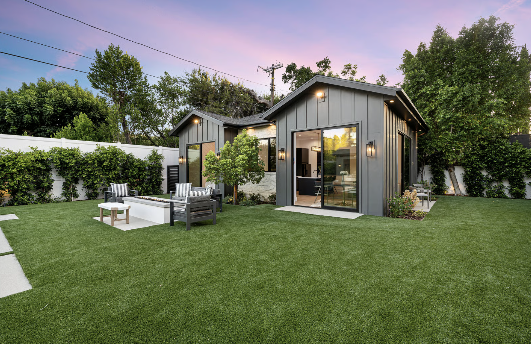 Modern backyard with dark gray siding house featuring large glass sliding doors, well-maintained green lawn, outdoor seating area with cushioned chairs around a rectangular fire pit, small trees, and tall hedges along a white fence at sunset.