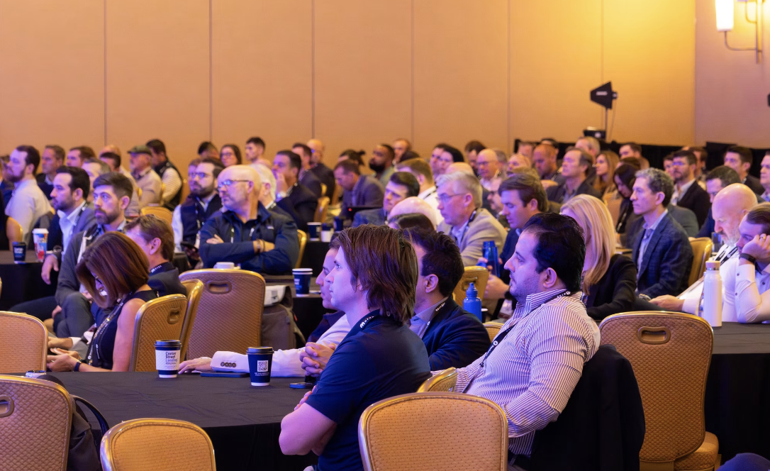 Audience of men and women seated in rows of chairs attentively listening to a speaker or presentation at a conference or seminar.