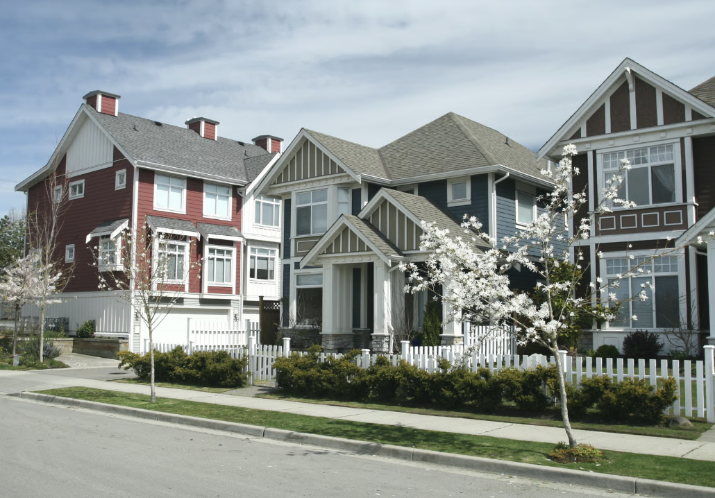 Three neighboring modern suburban houses with white picket fences, manicured lawns, bushes, and blossoming small trees lining a quiet street under a partly cloudy sky.