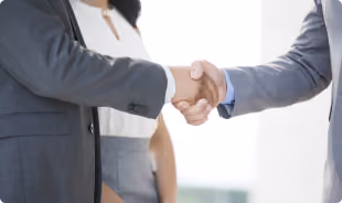 Two businesspeople shaking hands in a professional setting with a blurred person in the background.