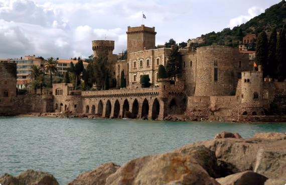 Historic stone fortress with turrets and arched wall along a body of water, with rocks in the foreground and cloudy sky.