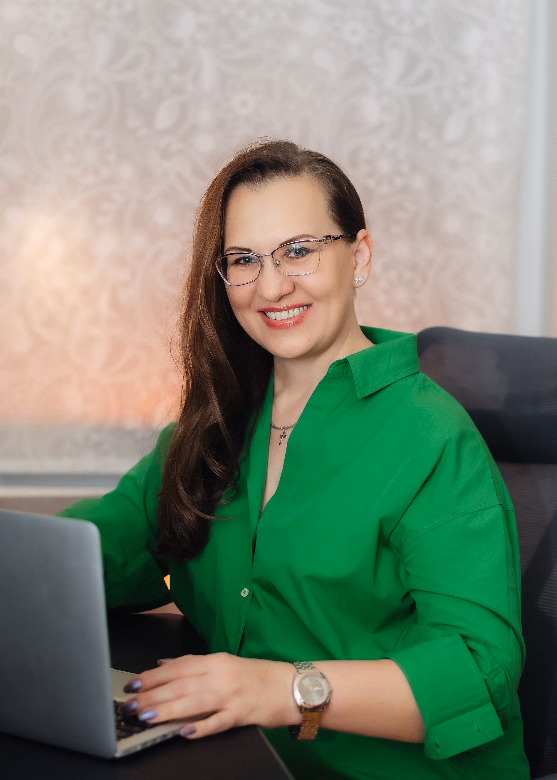 Frau mit langen braunen Haaren, die eine grüne Bluse trägt, sitzt an einem Schreibtisch und arbeitet an einem Laptop, dabei lächelt sie freundlich in die Kamera.
