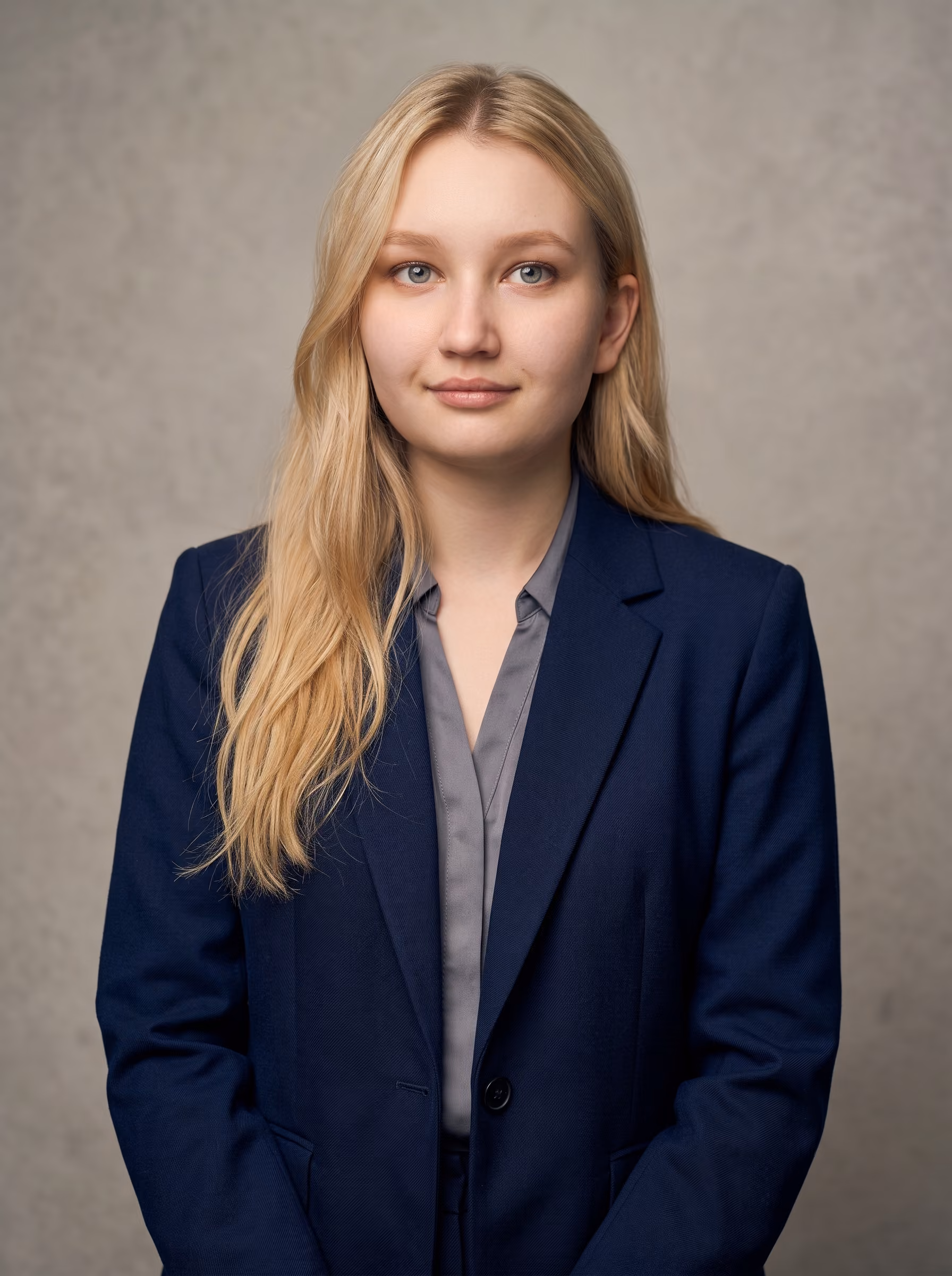 Junge Frau mit langen blonden Haaren trägt eine dunkelblaue Anzugjacke und ein graues Hemd, vor einem neutralen grauen Hintergrund.