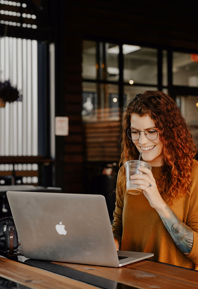 Smiling woman with red curly hair drinking an iced coffee while working on her laptop in a café