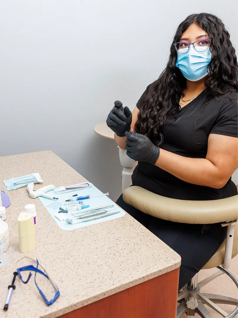 Woman dental professional wearing face mask and gloves sitting beside a dental tools tray.