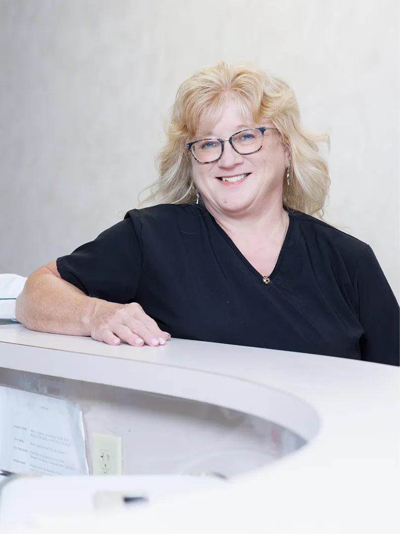 Smiling woman with blonde hair and glasses leaning on a white counter in a light room.