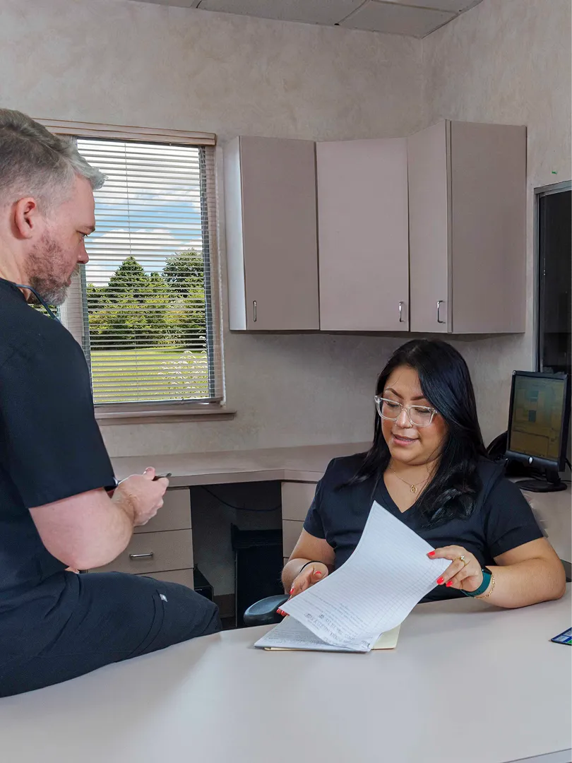 Two medical professionals in black scrubs reviewing documents in a bright office with a window.
