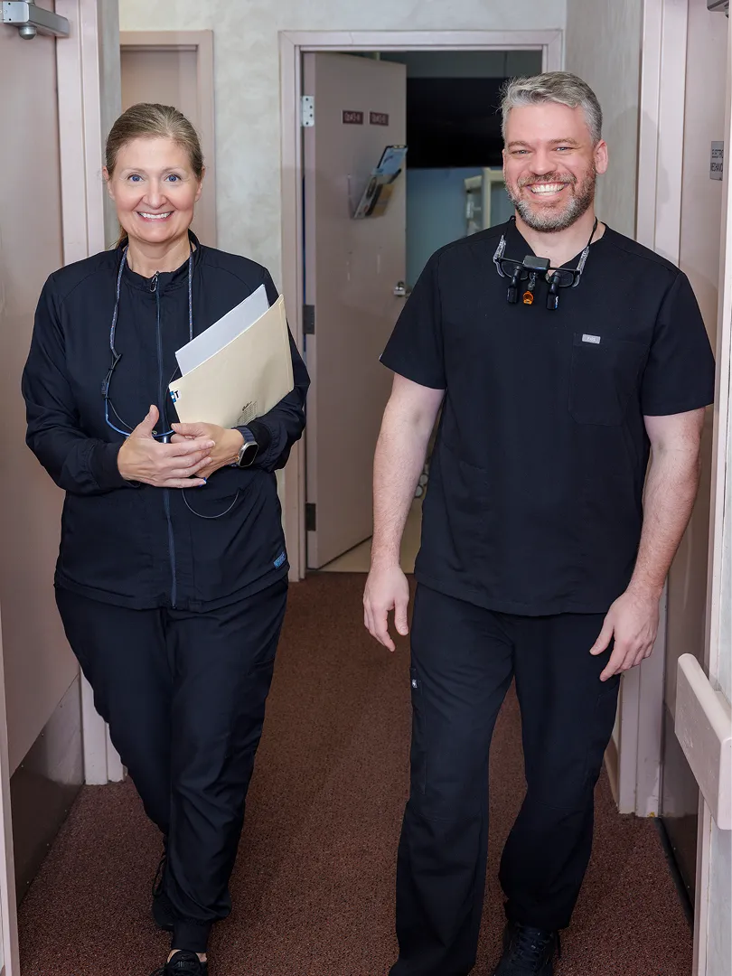 Two smiling dental professionals walking in a hallway wearing black scrubs with dental loupes.