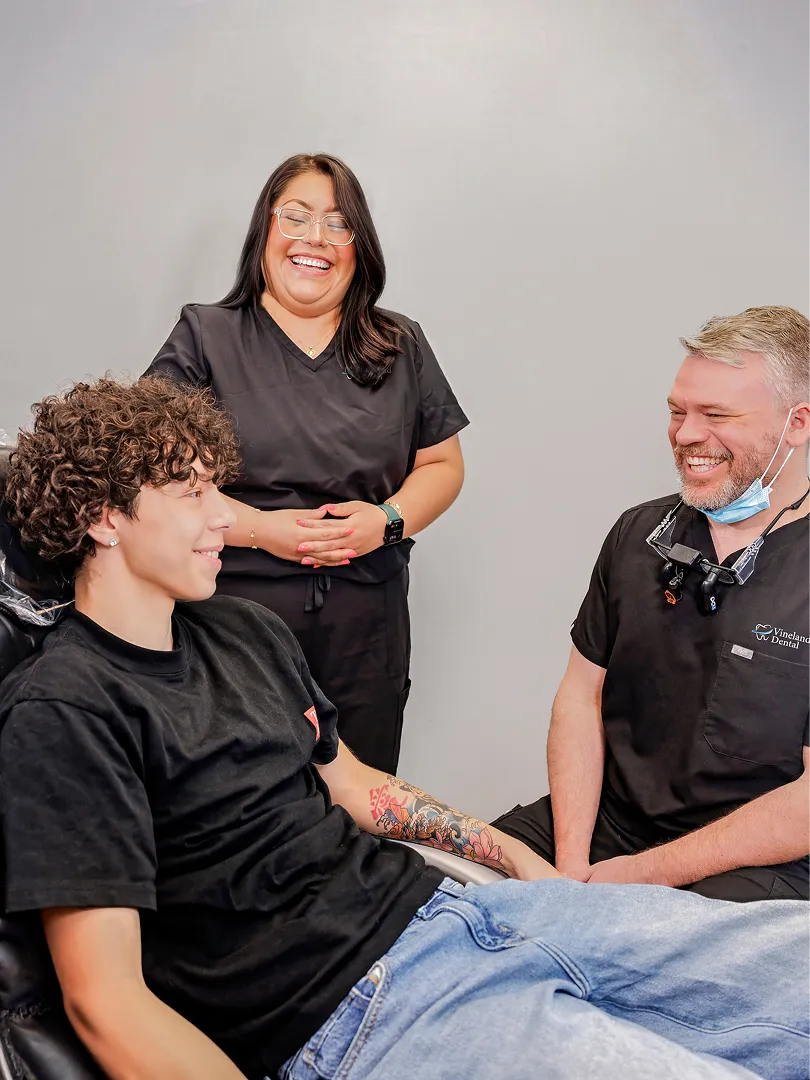 Young man sitting in dental chair smiling with two dental staff, all appearing happy and relaxed.