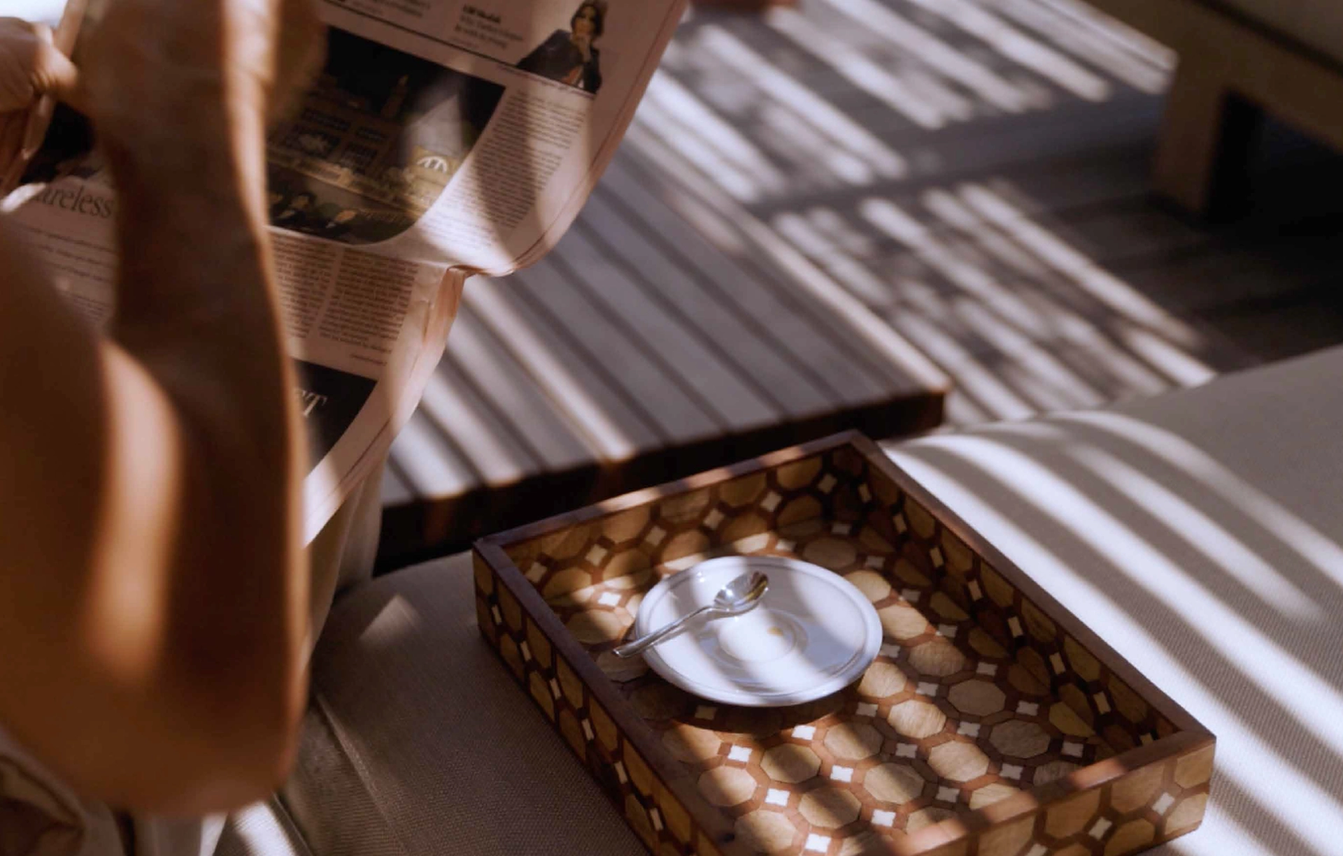 A person reading a newspaper next to a decorative wooden tray with a white saucer and spoon on a sunlit surface.