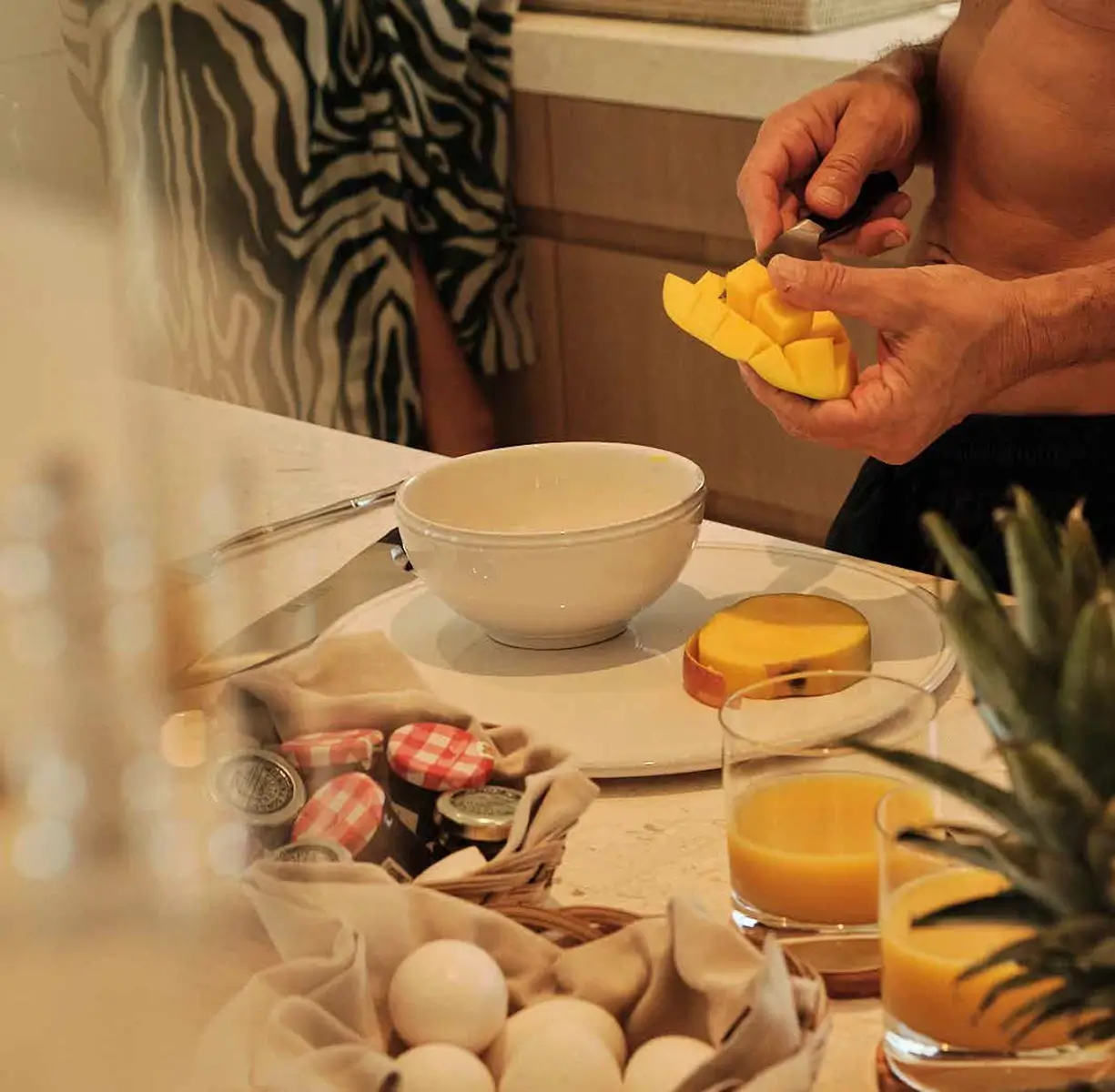 Person carving a diced mango half over a white bowl on a kitchen counter with juice and eggs nearby.