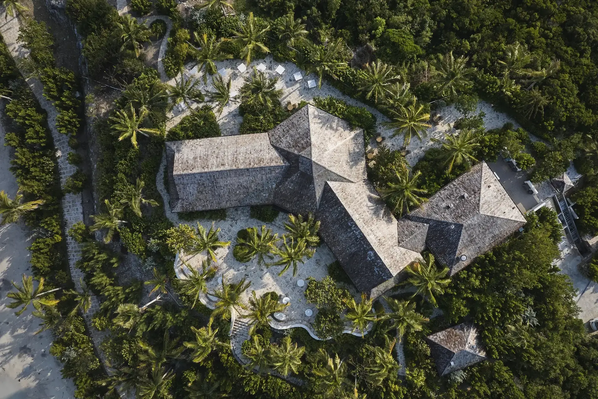 Aerial view of a large house with gray roofs surrounded by lush green trees and palm trees.
