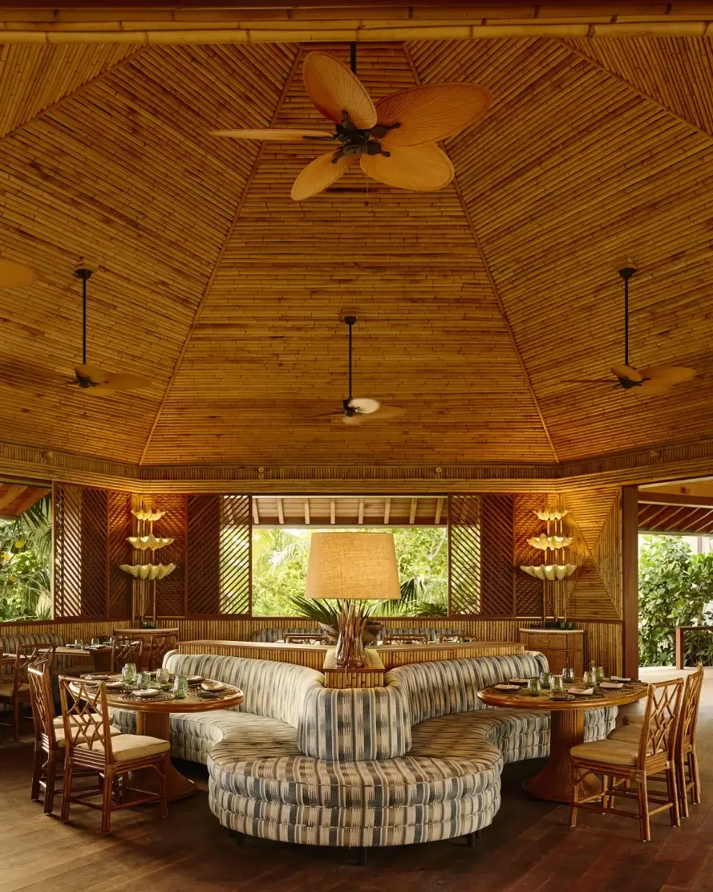 Indoor tropical restaurant with bamboo ceiling, palm leaf fans, round booths, and wooden tables and chairs.