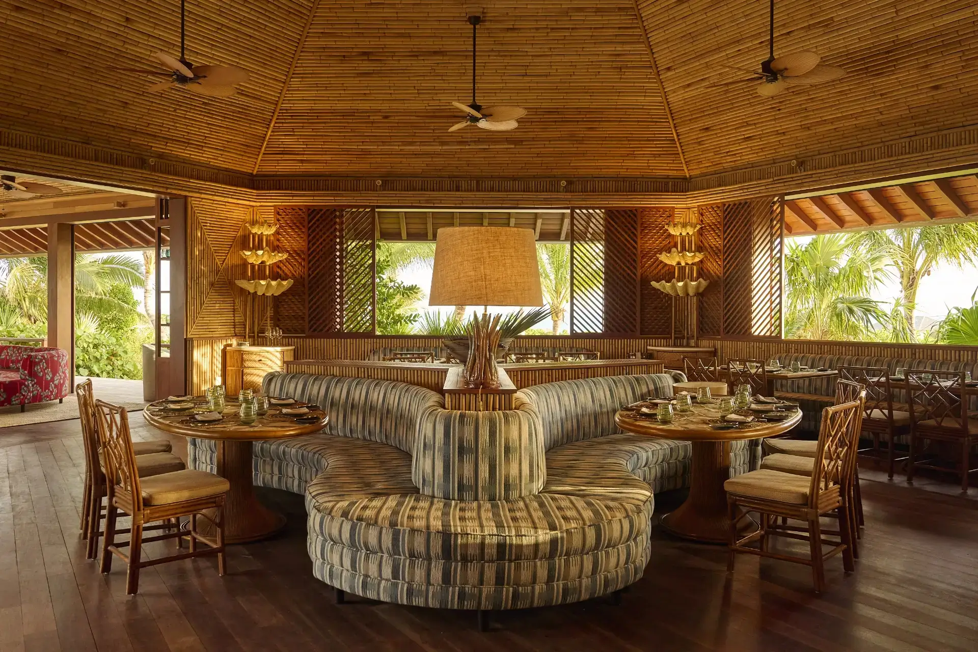 Cozy tropical restaurant interior with bamboo ceiling, round booths, wooden chairs, and large central lamp.