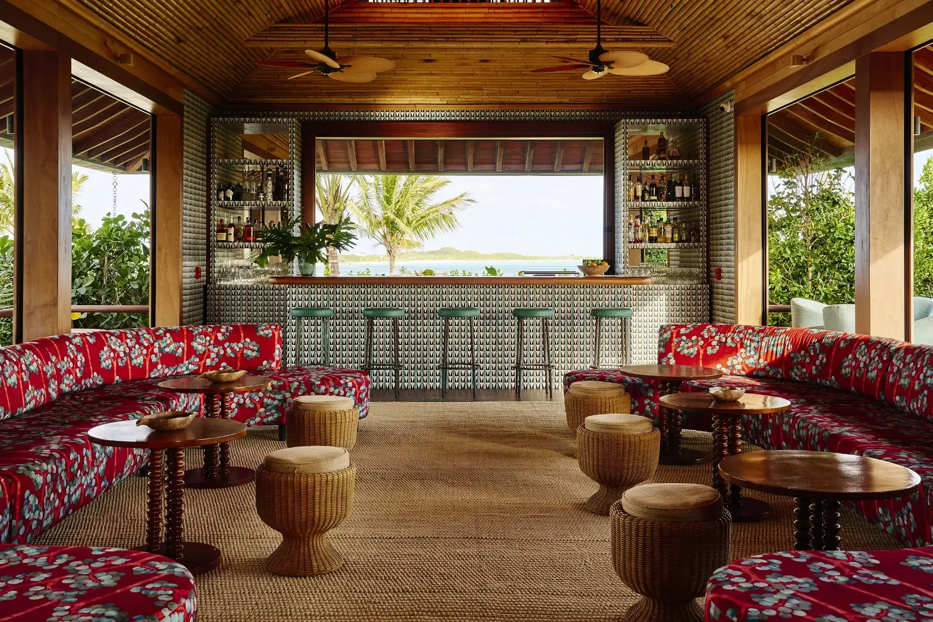 Cozy tropical bar with red floral couches, wicker stools, wooden tables, and ocean view through window.
