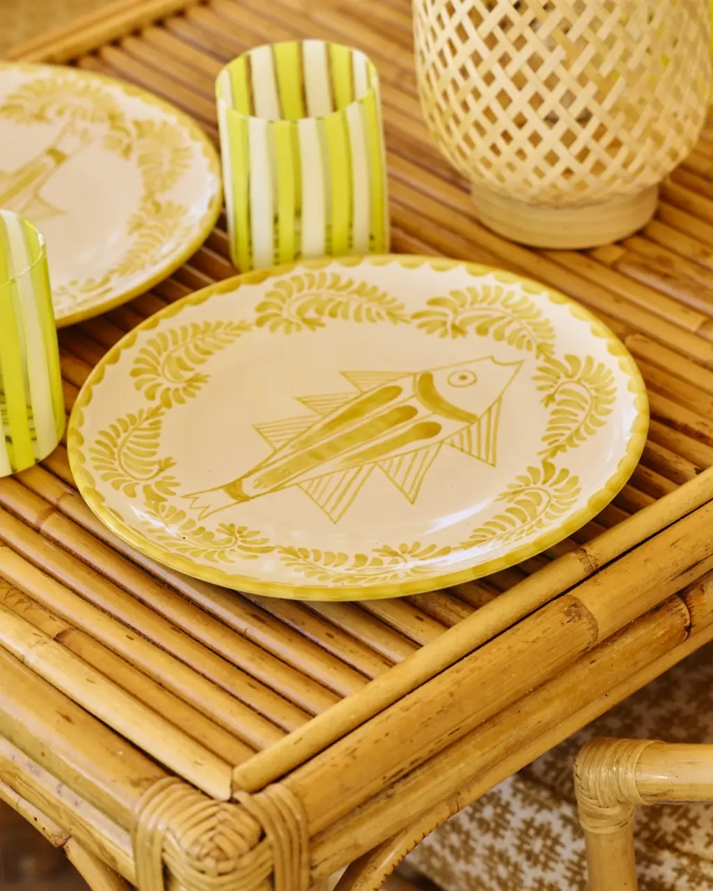 Yellow fish-decorated plate on a bamboo table beside two striped yellow-green cups and a woven lantern.