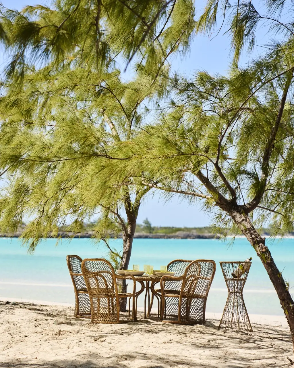 Wicker chairs and table under trees on sandy beach by turquoise sea with wine bottle holder nearby.