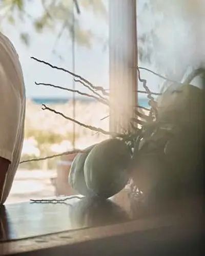 Green coconuts with stems resting on a wooden surface near a window with a blurred outdoor beach scene in the background.