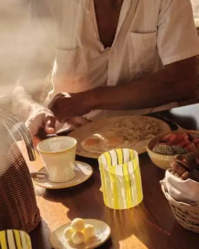 Person in a white shirt sitting at a wooden table with a cup of coffee, a yellow-striped glass, a plate with round white treats, and baskets containing fruits and bread, with warm sunlight streaming in.