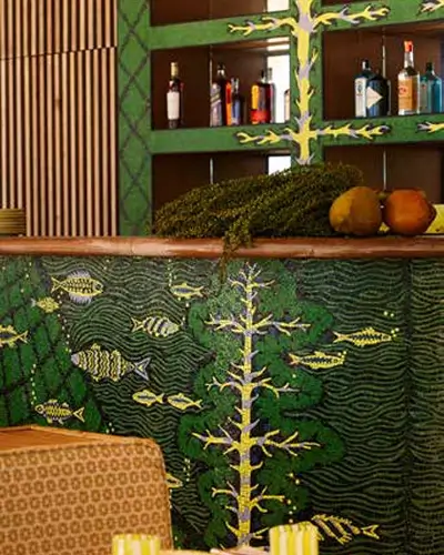 Close-up of a bar decorated with a green aquatic-themed mural featuring stylized yellow fish and plant-like patterns, with bottles of liquor on shelves behind and a cozy chair in the foreground.