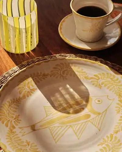 Table setting with a decorative plate featuring a yellow fish illustration and floral border, a cup of black coffee in a beige cup and saucer, and a yellow striped glass casting a shadow on the plate.