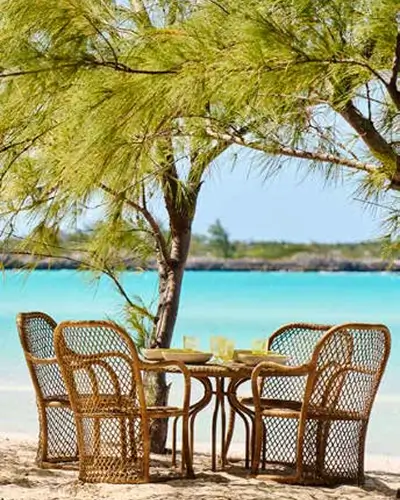 Four wicker chairs around a wooden table set with glasses and plates under a tree on a sandy beach with turquoise water and a clear sky in the background.