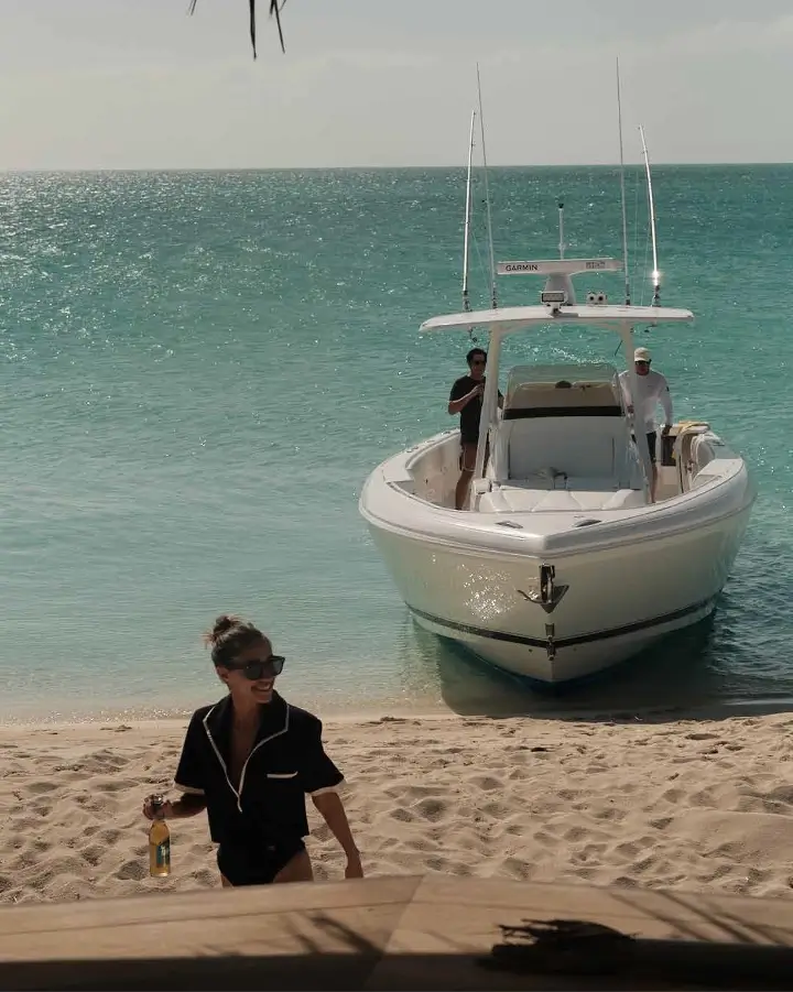 A woman in dark clothing and sunglasses walking on a sandy beach holding a bottle, with a white motorboat anchored near the shore and two people standing on the boat, turquoise ocean water in the background.