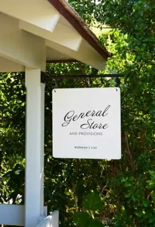 White hanging sign on a black metal bracket reading 'General Store and Provisions Norman's Cay' in front of green foliage and part of a white building roof.