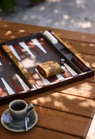 A backgammon board set on a wooden table outdoors with dice and a doubling cube on the board, along with a ceramic cup and saucer with a spoon on the side.