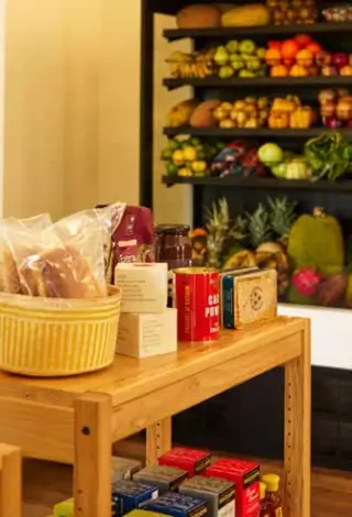 Wooden table in a general store displaying jars, canned goods, and baskets of packaged bread, with shelves of fresh fruits and vegetables in the background.