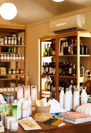 Interior view of a small general store with wooden shelves stocked with jars, bottles, and various products, a table in the foreground displaying skincare items, a woven basket, books, and reusable water bottles under a ceiling with round white hanging lights.
