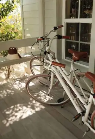 Two white bicycles with brown seats and handles parked on a wooden porch next to a white railing and a window with multiple panes, with sunlight and shadows on the floor.