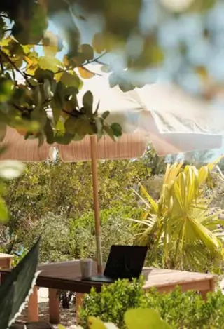 Outdoor wooden table with a laptop and drink under a beige umbrella surrounded by green plants and trees.