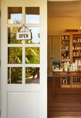 White glass-paneled door with an "OPEN" sign, partially revealing an interior of a wooden general store with shelves and a table displaying various items.
