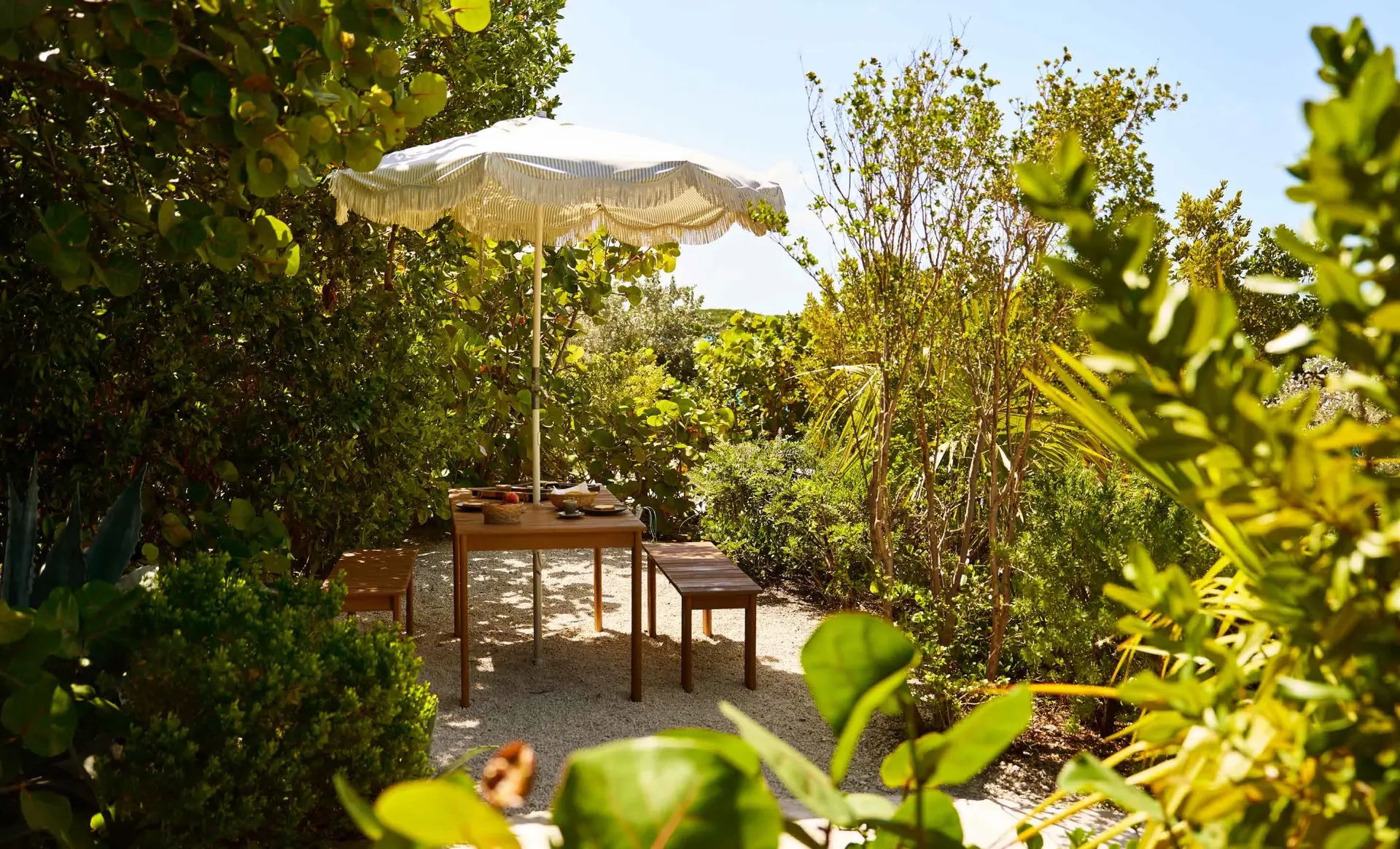 Outdoor seating area with a wooden table and two benches under a fringed beige umbrella, surrounded by lush green foliage and shrubs on a sunny day.