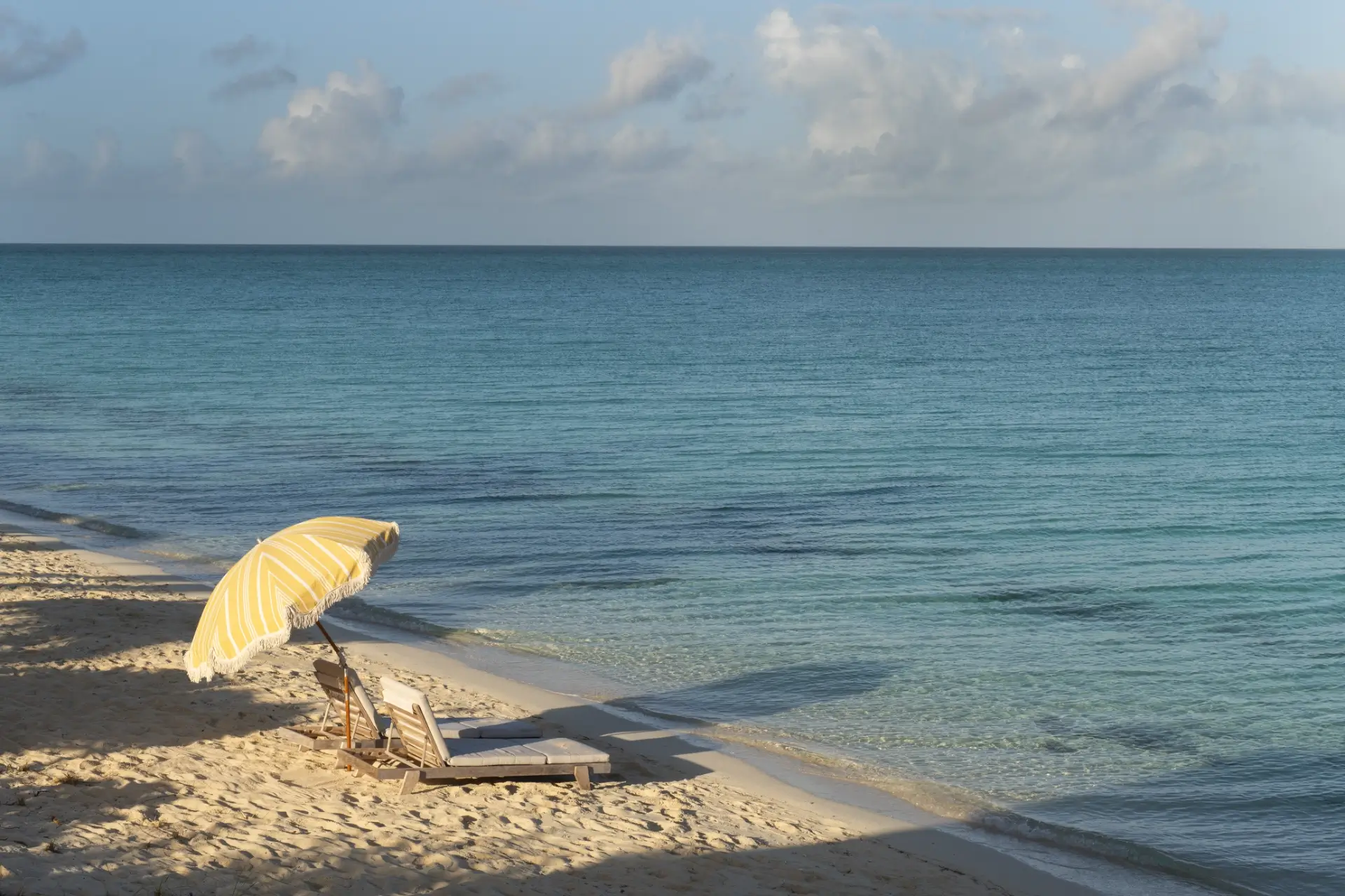 Two wooden lounge chairs with cushions under a yellow striped umbrella on a sandy beach next to calm blue ocean water under a partly cloudy sky.