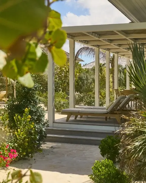 Two wooden lounge chairs with cushions under a covered patio surrounded by lush green plants and trees, with a clear sky in the background.