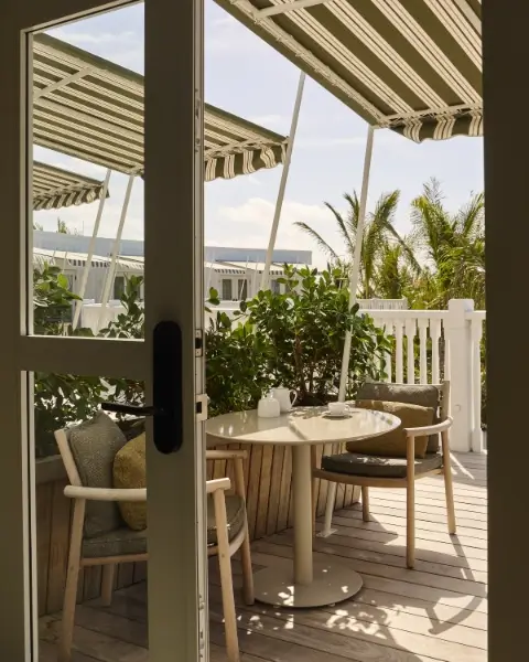 View through a glass door of a cozy outdoor patio with a round table and two wooden chairs, green plants, white fence, striped awnings, and palm trees in the background under a clear sky.