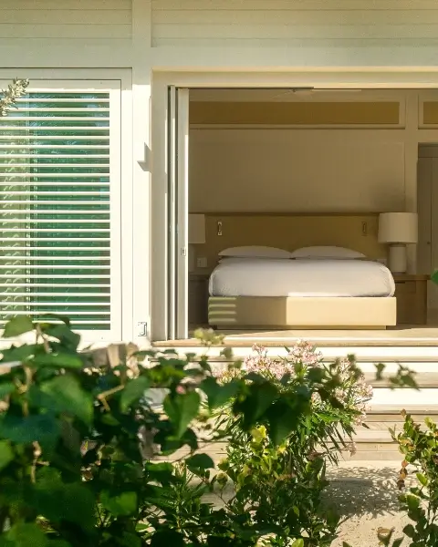 View of a neatly made bed with white bedding inside a room through a large open window, with green plants and flowers in the foreground outside.