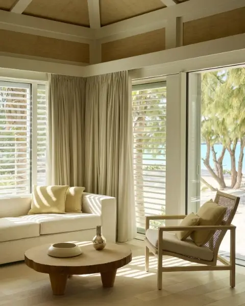 Bright living room with a white sofa and beige cushions, a wooden round coffee table with decorative items, a beige armchair with cushions, large windows and glass doors showing trees and beach outside.