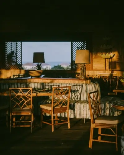 Cozy indoor seating area with wooden chairs and cushioned bench seats under warm lighting, framed by a large window showing an outdoor view.